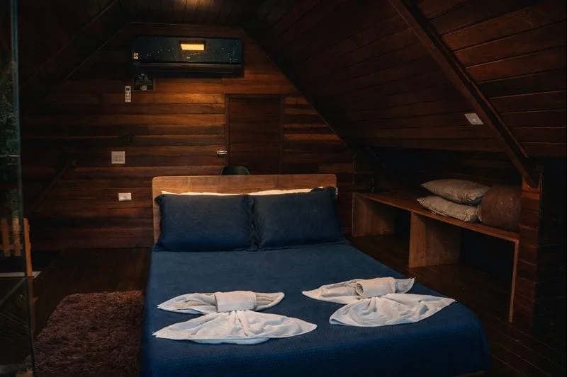 Mezanino do chalé com cama de casal arrumada em tons de azul, toalhas decorativas e ar-condicionado