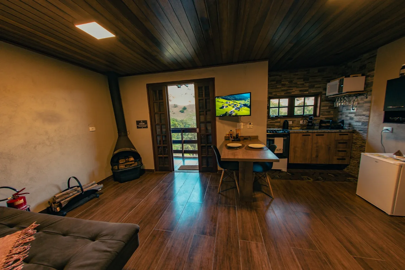 Sala e cozinha integradas do chalé, com lareira, mesa de jantar e porta aberta para a varanda
